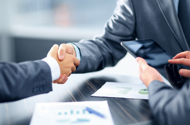 Two people in business suits shaking hands across a table with business documents and a tablet.