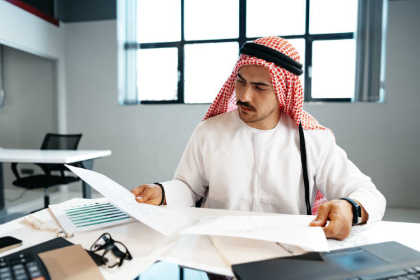 Man in traditional attire reviewing documents at a desk, analyzing Ajman Free Zone license fees.