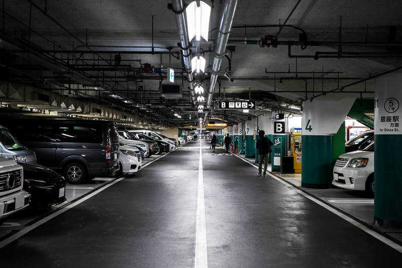 A dimly lit, underground parking garage with parked cars on both sides and people walking towards an elevator entrance.