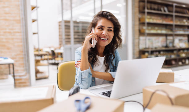 A small business owner looks happy while talking on the phone and working on a laptop in her office.